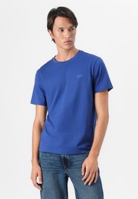T-shirt in cotone blu con colletto a giro e maniche corte. Logo discreto sul petto. Il modello lo indossa con jeans in denim blu.