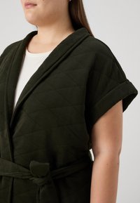 Tamsiai žalia, ketvirtine danga suknelė su apykakle, suvyniotos rankovės ir juosta. Minkšta tekstūra ir deimanto rašto detalės.