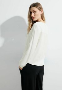 Weiße Bluse mit langen Ärmeln, weicher Textur und leicht drapiertem Rücken, kombiniert mit schwarzen Hosen. Verleiht einen schlichten, klassischen Look.