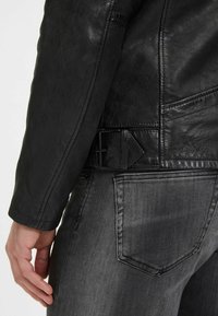 Zwarte leren jas met gedetailleerde stiksels en een ruitvormige gesp op de taille. Grijze denim broek met een zichtbare achterzak.