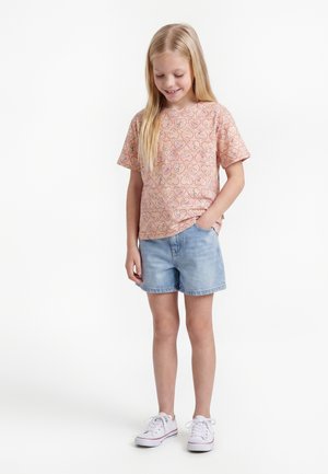 Jong meisje met lang blond haar, gekleed in een roze shirt met korte mouwen en een patroon, lichtblauwe spijkershorts en witte sneakers, staand en glimlachend.