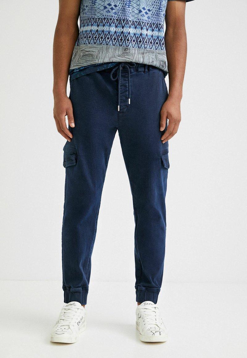 Pantalon cargo bleu marine avec poignets élastiques, ceinture à cordon de serrage et deux poches latérales. Associé à une chemise à manches courtes bleue à motifs et à des baskets blanches.