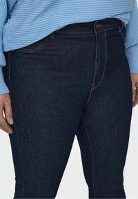 Person som bär mörkblå skinny jeans och en ljusblå ribbad tröja med uppvikt ärmar. Händerna hänger avslappnat längs sidorna.