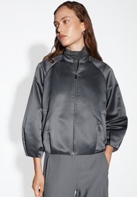 Giacca bomber in satin grigio con chiusura a zip, maniche raglan e una texture liscia. Indossata sopra un dolcevita aderente, abbinata a pantaloni grigi.