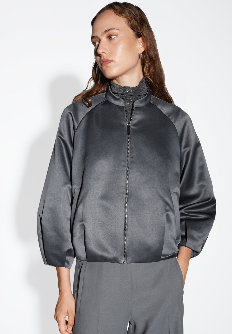 Giacca bomber in satin grigio con chiusura a zip, maniche raglan e una texture liscia. Indossata sopra un dolcevita aderente, abbinata a pantaloni grigi.