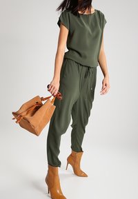 Combinaison verte olive à manches courtes, avec une taille à cordon, associée à un sac à main en suède marron et des bottes à talons assorties, texture lisse.