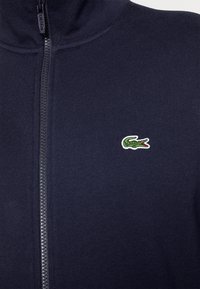 Bunda na zip z tmavě modré látky s jemnou texturou. Na levé straně hrudi je vyšívané zeleno-červené logo krokodýla.