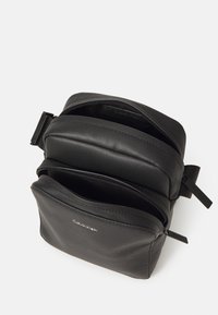 Borsa in pelle nera con finitura texturizzata, dotata di due scomparti con zip e un logo. Forma compatta e rettangolare con tracolla regolabile.