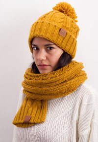 Mustard yellow knit beanie with a pom-pom and matching scarf, both featuring thick, textured patterns and a leather logo accent.