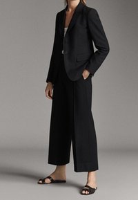 Zwarte getailleerde blazer met één knoop, gecombineerd met wijde zwarte broeken. Gladde stof met minimale details, aangevuld met platte zwarte sandalen.