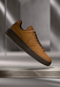 Mustard yellow leather sneaker with a smooth texture, brown rubber sole, perforated toe, and logo detail on the heel.