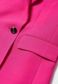 Gros plan d'un blazer rose vif avec un bouton noir et une poche à rabat sur un tissu lisse.