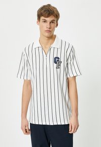 Valkoinen lyhythihainen polo paita, jossa on tummansinisiä pystysuoria raitoja, vetoketjukaulus ja vasemmalla puolella tummansinisestä ja harmaasta brodeerattu "CF"-logo.