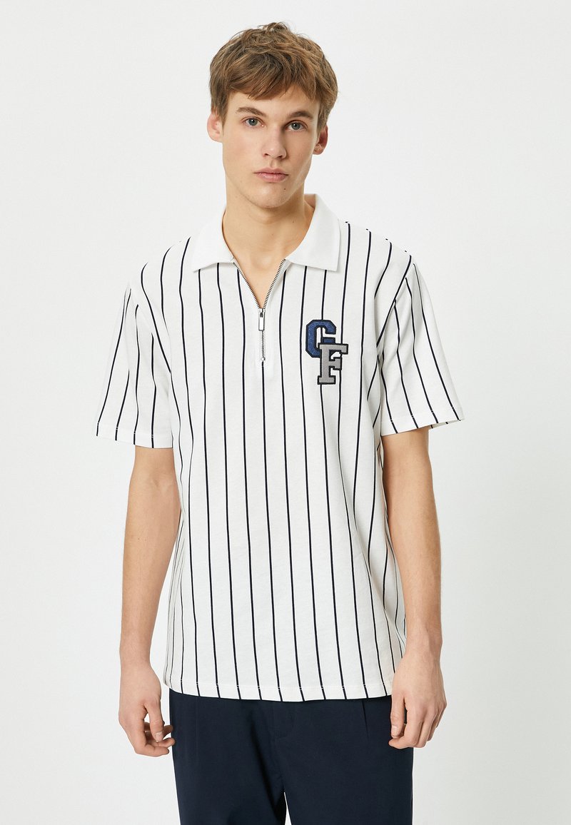 Valkoinen lyhythihainen polo paita, jossa on tummansinisiä pystysuoria raitoja, vetoketjukaulus ja vasemmalla puolella tummansinisestä ja harmaasta brodeerattu "CF"-logo.