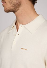 Polo shirt couleur crème avec un tissu texturé, présentant un col et trois petits motifs de nounours brodés marron sur la poitrine.