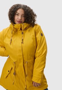 Yellow waterproof jacket with drawstring hood, zip and snap closure, elastic cuffs, and side pockets. Smooth texture with a slightly glossy finish.