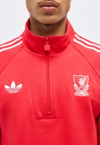 Noor mees, kes kannab punast lukuga spordijopet, mille rinnal on valged Adidas ja Liverpooli jalgpalliklubi logod.