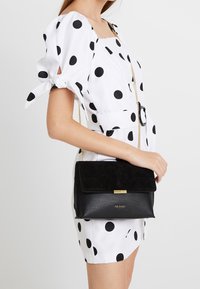 Černá kožená taška s semišovým klopou, s designovými zlatými akcenty. Taška má texturovaný povrch a řetízkový popruh na nošení.