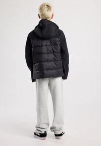 Mustahoodinen takki, jossa on paksu eristys selässä, harmaat verkkarit ja mustat lenkkarit. Materiaalit näyttävät sileiltä ja mattaisilta.