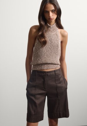 Donna che indossa un top beige lavorato a maglia senza maniche con scollo halter e pantaloncini marrone scuro a vita alta e lunghezza al ginocchio con le mani nelle tasche.