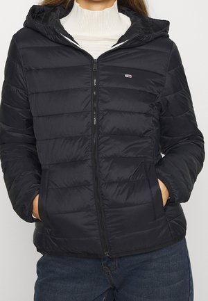 Kvinde iført en sort quiltet lynlåstrøje med hætte, hænderne i lommerne, og en hvid ribbet rullekraveundertrøje nedenunder.