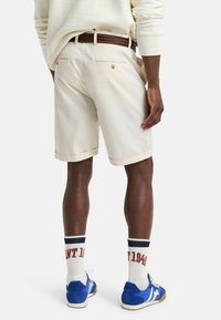 Beige katoenen shorts met opgerolde mouwen, gecombineerd met blauwe sneakers en witte sokken met rode letters. Bruine gevlochten riem zichtbaar.