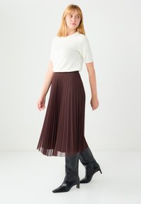 Jupe midi plissée marron à couches transparentes, associée à un haut blanc à manches courtes et des bottes à talons noires. Design simple, texture lisse.