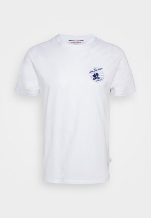 Persona che indossa una T-shirt bianca a maniche corte con logo, jeans azzurri a gamba dritta e scarpe da ginnastica arancioni e bianche, in piedi con una mano in tasca.