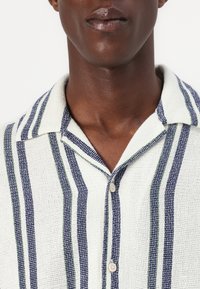 Camisa de manga corta en tejido entrelazado con fondo blanco, rayas verticales azules y verdes, tapeta de botones y un diseño de cuello clásico.