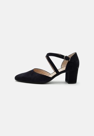 Chaussure noire en suède à bout fermé, talon carré, avec des brides croisées à la cheville et une boucle argentée sur fond blanc.