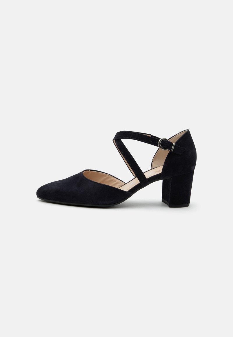 Chaussure noire en suède à bout fermé, talon carré, avec des brides croisées à la cheville et une boucle argentée sur fond blanc.