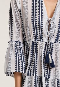 Blau-weiß gemustertes Kleid mit ausladenden Ärmeln, V-Ausschnitt, Krawattenverschluss und Quastenakzenten. Das Material scheint leicht und strukturiert zu sein.