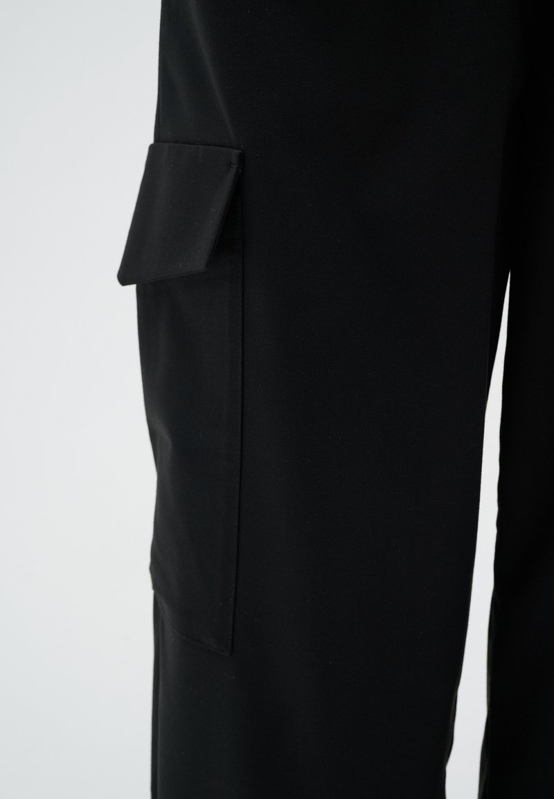 Schwarze Stoffhose mit einer seitlichen Klappentasche am Oberschenkel, vor einem schlichten hellen Hintergrund gezeigt.