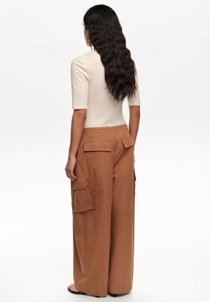 Pikkade laineliste tumedate juustega naine, kes kannab beežikat lühikeste varrukatega topi ning kõrge vöökohaga laia säärega pruune pükse, millel on suured klappidega taskud, vaade tagant.