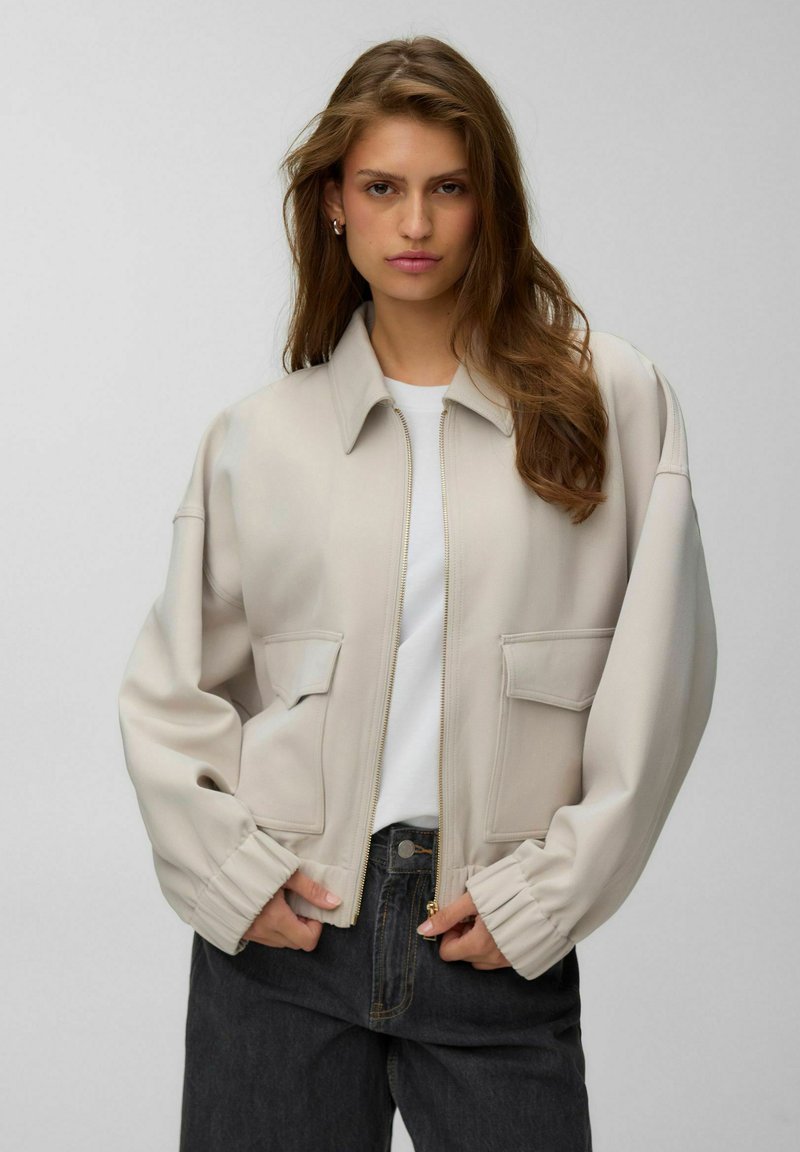Femme aux longs cheveux bruns portant une veste beige zippée par-dessus une chemise blanche et un jean foncé, debout les mains près des poches de la veste.