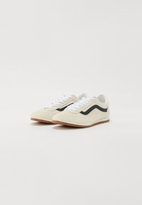 Beige Wildlederschuhe mit weißen Schnürsenkeln und schwarzem Streifenakzent. Sie verfügen über eine Gummisohle mit strukturierter Oberfläche für besseren Halt.