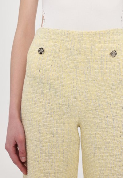 Keltaiset tweed-housut, joissa on rakenteellinen pinta, kahdella etutaskulla ja mustilla koristepainikkeilla kummallakin puolella.