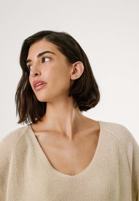 Beige neulepaita, jossa on avara V-pääntie ja ribbikuviointi, tyyliin kuuluen pudotetut olkapäät ja rento istuvuus, taustalla yksinkertainen tausta.