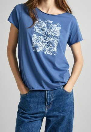 Frau trägt blaues Kurzarmshirt mit weißem Blumen- und Schriftmuster, leicht in blaue Jeans gesteckt, Hände in den Taschen.