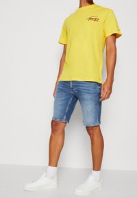 T-shirt di cotone giallo con logo nero, abbinato a pantaloni corti in denim blu. Il modello indossa sneaker bianche e calzini alla caviglia. Design minimalista.