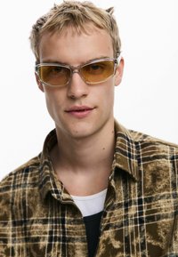 Lunettes de soleil avec des verres teintés jaunes et une monture transparente, portées avec une chemise en flanelle à carreaux marron sur un haut noir.
