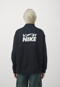 Černá oversized mikina s velkým bílým logem "NIKE" na zadní straně, s hladkým povrchem a žebrovanými lemy na rukávech a spodním okraji.
