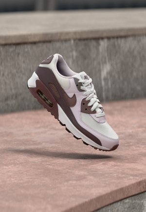Biały i liliowy but Nike Air Max unoszący się nad kamienną powierzchnią, na tle szarej kamiennej ściany.