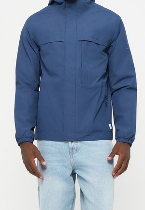 Mann trägt eine blaue Kapuzenjacke mit elastischen Bündchen und hellblauen Jeans und steht vor einem einfarbigen Hintergrund.