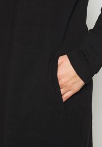 Melnā adīta kleita ar garām piedurknēm, ar sānu kabatām, gludu faktūru un slaidu piegriezumu, izceļot roku kabatā.