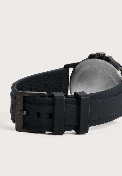 Gros plan sur le bracelet en caoutchouc, la boucle et le fond de boîtier en métal d’une montre noire, sur un fond clair.