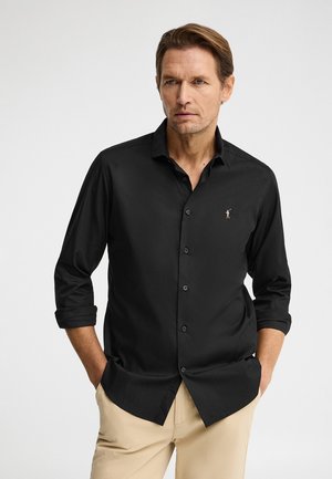 Mężczyzna w czarnej koszuli na guziki z podwiniętymi rękawami i beżowych spodniach, stojący z rękami w kieszeniach na jednolitym jasnym tle.