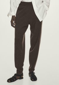 Un pantalon brun, ample et léger, avec une texture douce et une taille élastique, associé à des sandales noires à lanières.