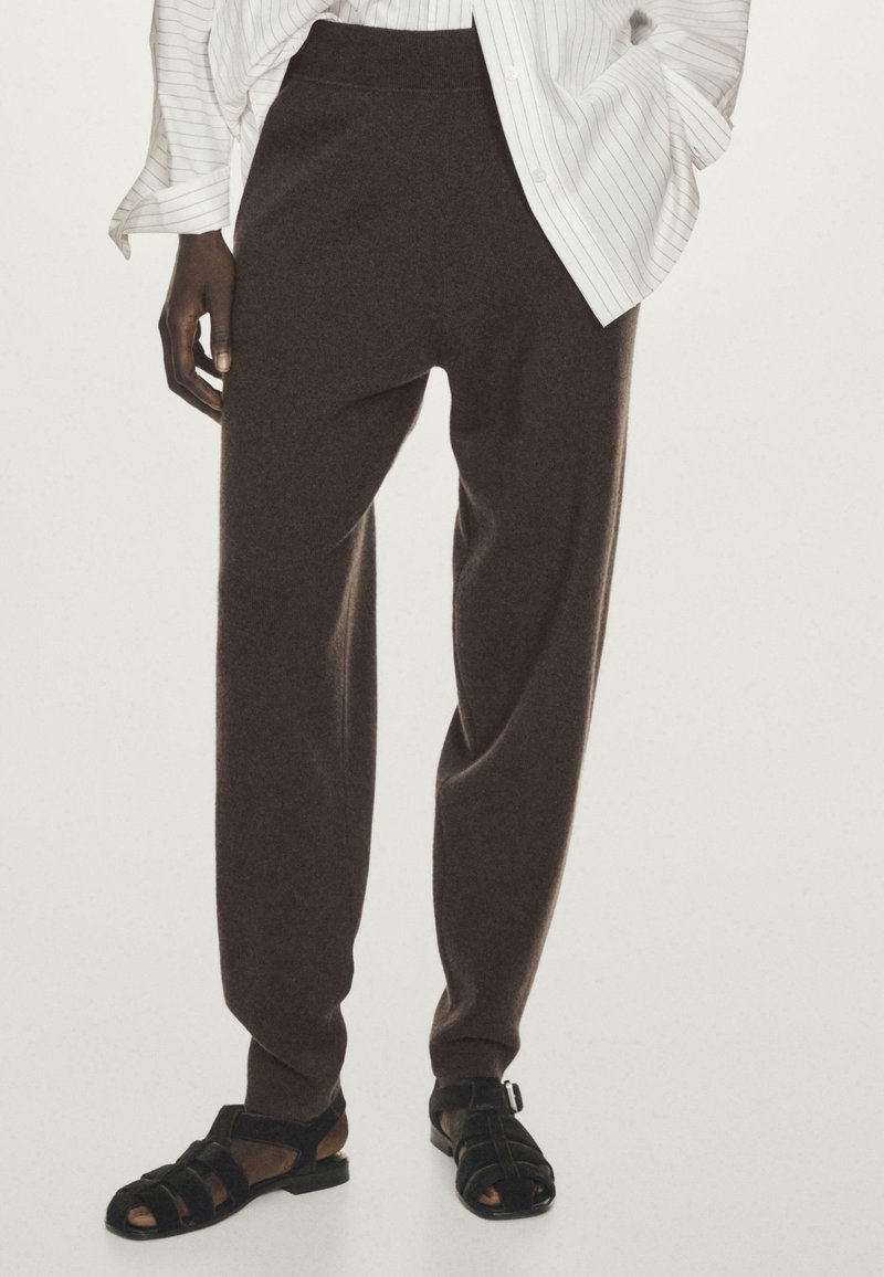 Un pantalon brun, ample et léger, avec une texture douce et une taille élastique, associé à des sandales noires à lanières.