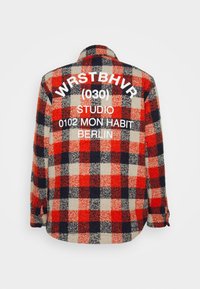 Camisa de flanela xadrez em vermelho, preto e bege, com grandes letras brancas nas costas: "WR$TBHVR (030) STUDIO 0102 MON HABIT BERLIN."